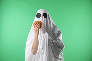 Photo of Creepy ghost. Person in white sheet with tasty hamburger on green background