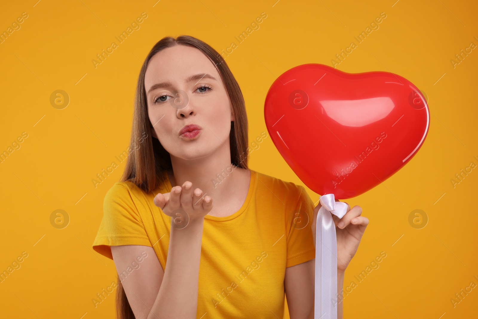 Photo of Young woman holding red heart shaped balloon and blowing kiss on yellow background