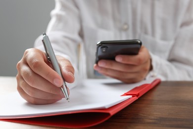 Photo of Woman with smartphone writing on sheet of paper in red folder at wooden table in office, closeup