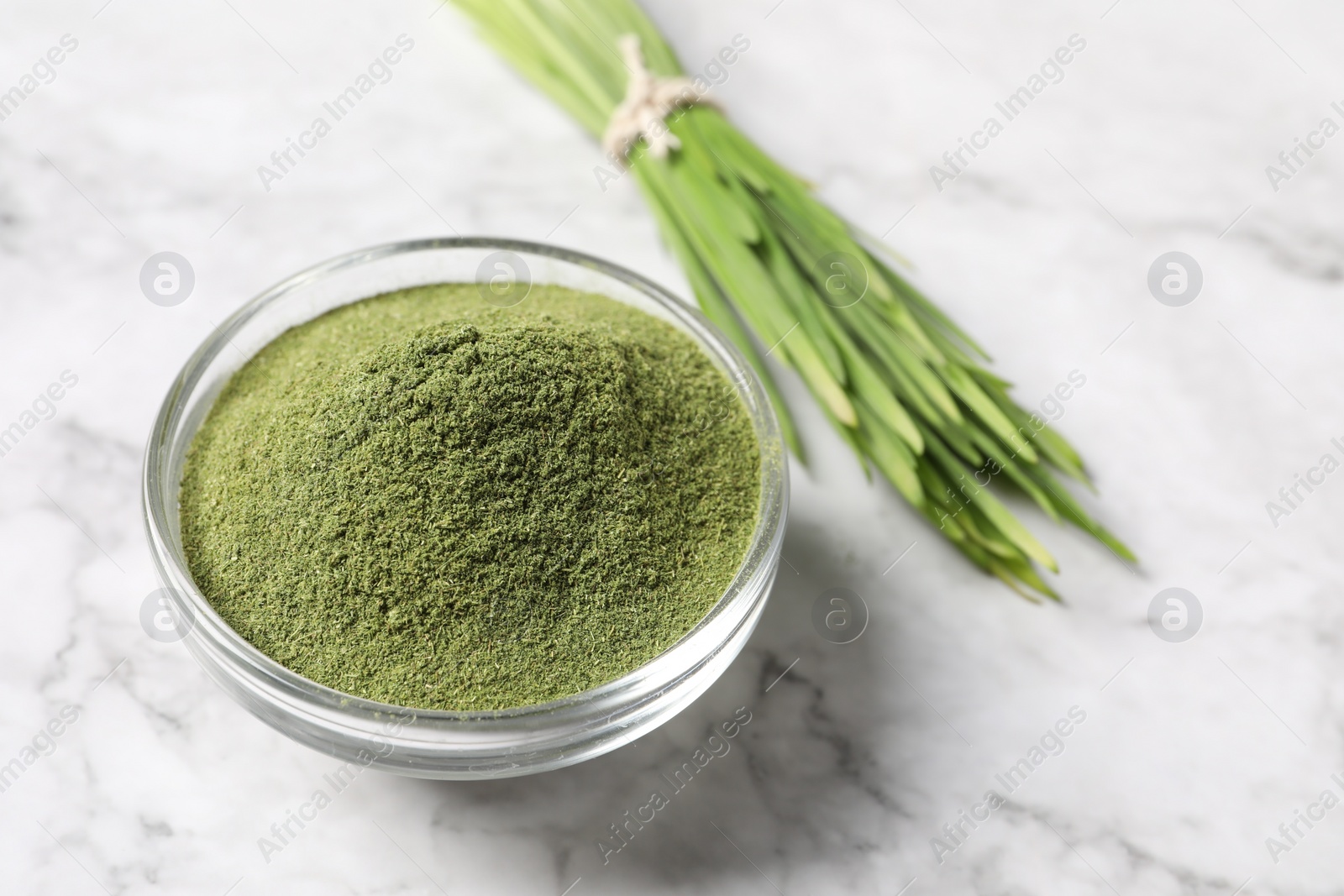 Photo of Wheat grass powder in glass bowl on white marble table, closeup