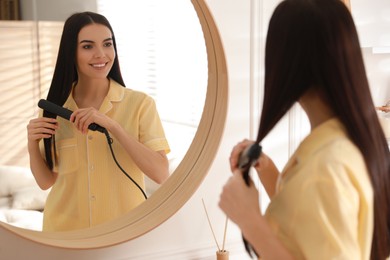 Photo of Young woman straightening hair near mirror at home. Morning routine