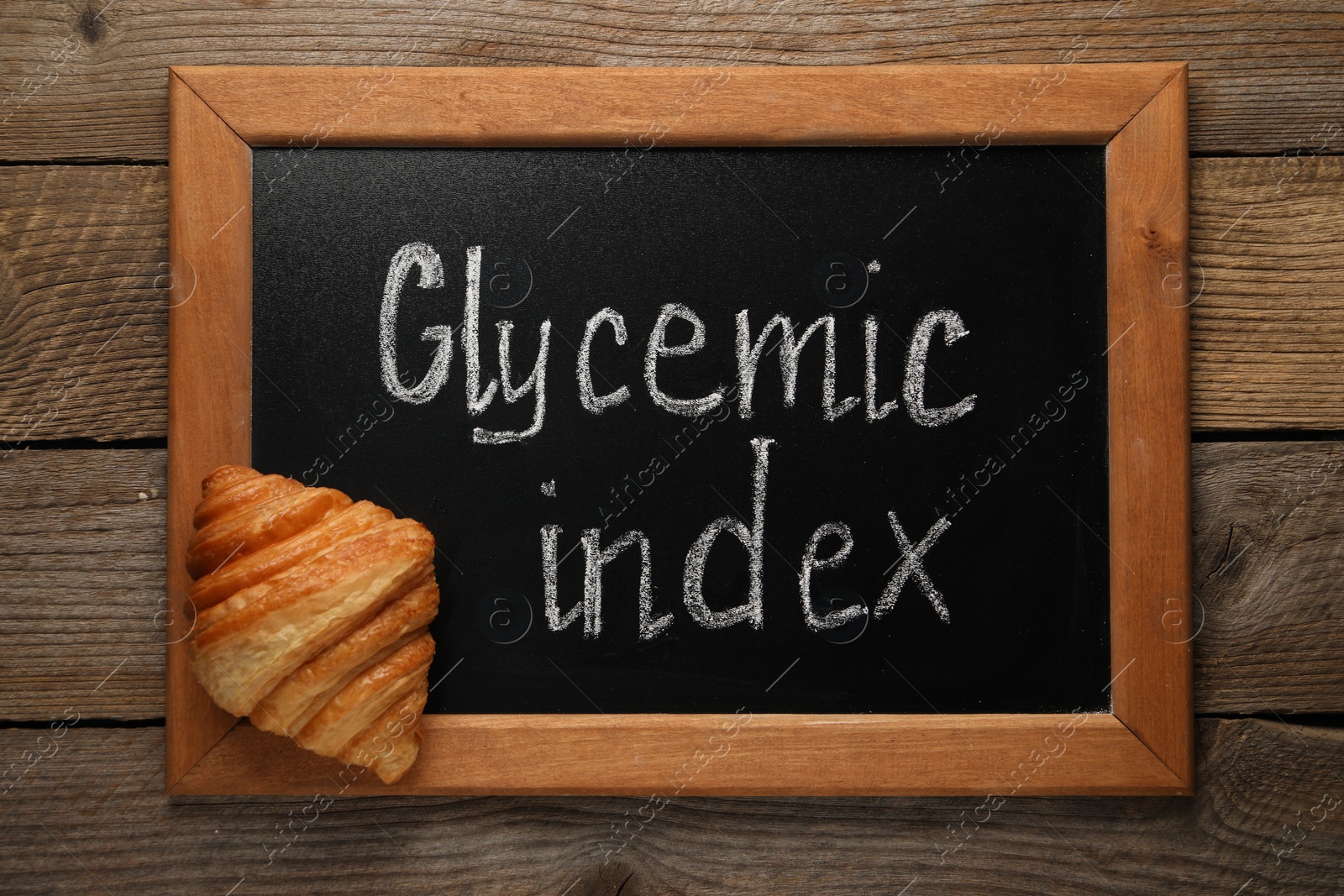 Photo of Small chalkboard with words Glycemic Index and croissant on wooden table, top view