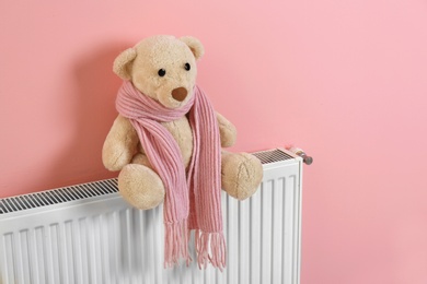 Photo of Teddy bear with knitted scarf on heating radiator near color wall