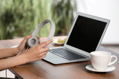 Photo of Woman holding headphones at wooden table with laptop, closeup