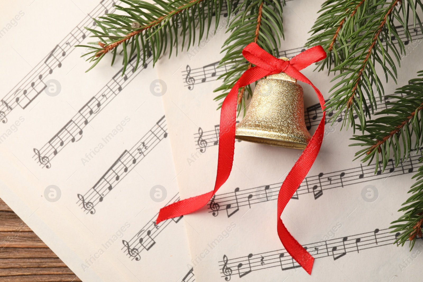 Photo of Golden shiny bell with red bow, fir branches and music sheets on wooden table, flat lay. Christmas decoration