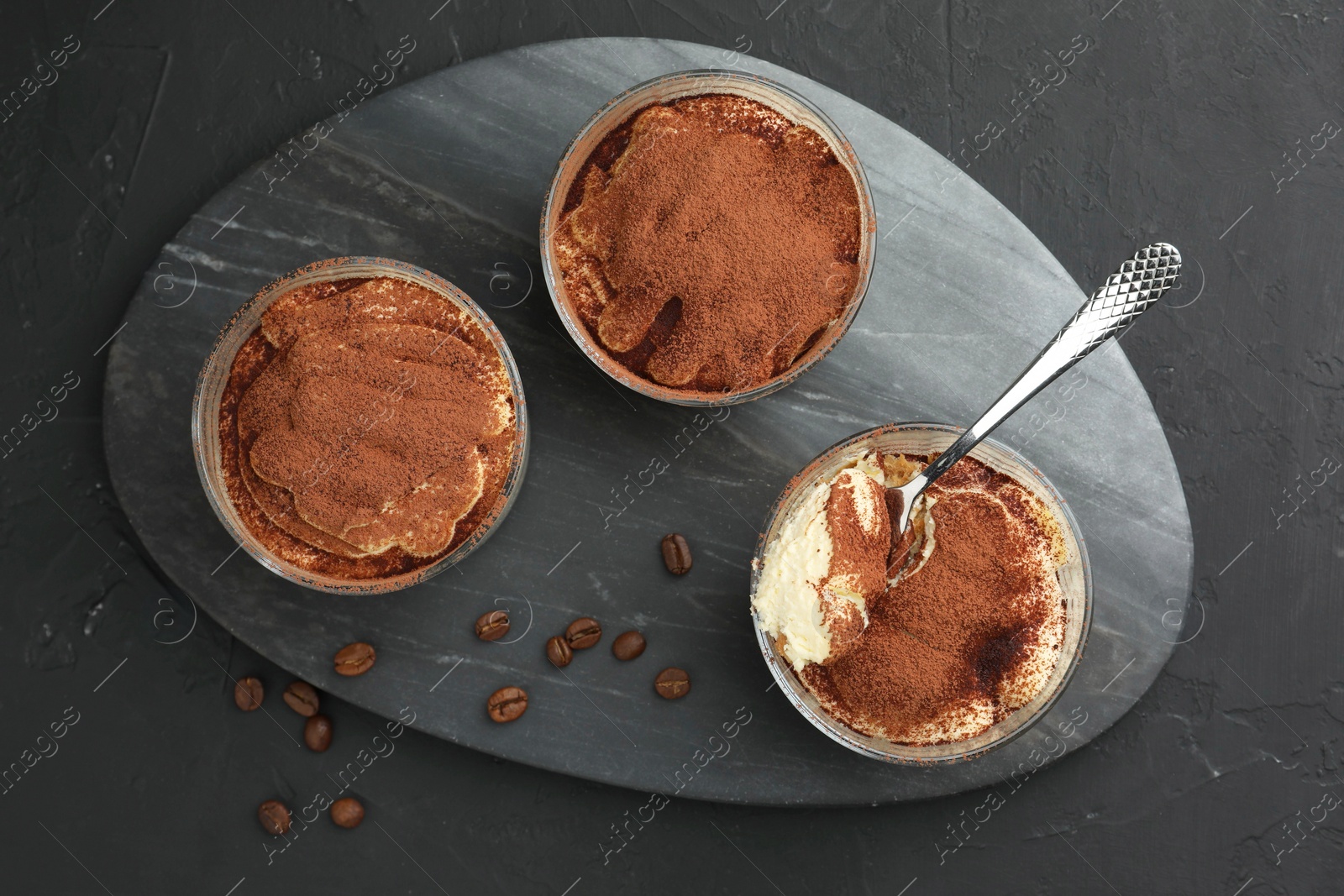 Photo of Delicious tiramisu in glasses, spoon and coffee beans on black table, top view