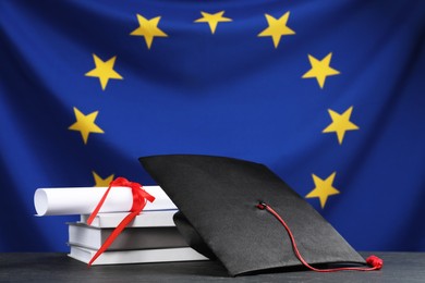 Photo of Graduation cap, diploma and books on black table against flag of European Union
