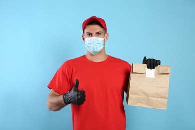 Courier in medical mask holding paper bag with takeaway food on light blue background. Delivery service during quarantine due to Covid-19 outbreak
