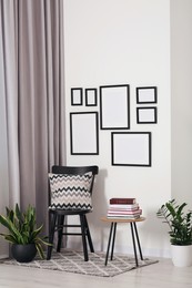 Photo of Empty frames hanging on white wall, wooden chair and table with books indoors