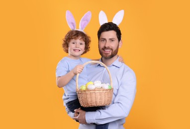 Photo of Happy father and son wearing cute bunny ears headbands on orange background. Boy holding Easter basket with painted eggs