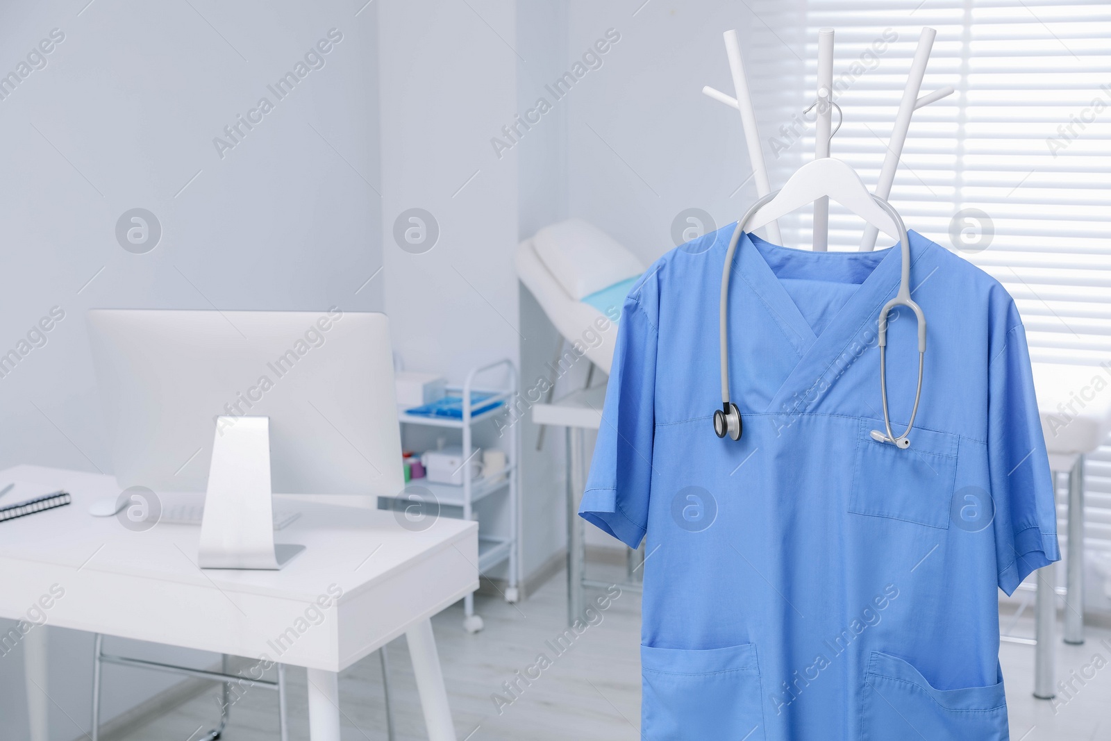 Photo of Blue medical uniform and stethoscope hanging on rack in clinic. Space for text