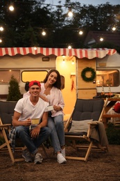 Happy couple with cups resting on deck chairs near motorhome. Camping season