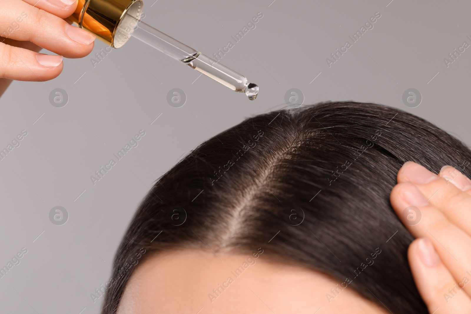 Photo of Woman applying essential oil onto hair roots on light grey background, closeup