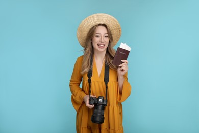 Happy young woman with camera, passport, ticket and hat on light blue background