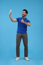 Smiling man taking selfie with smartphone and showing thumbs up on light blue background