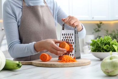 Photo of Woman grating fresh ripe carrot at kitchen table, closeup