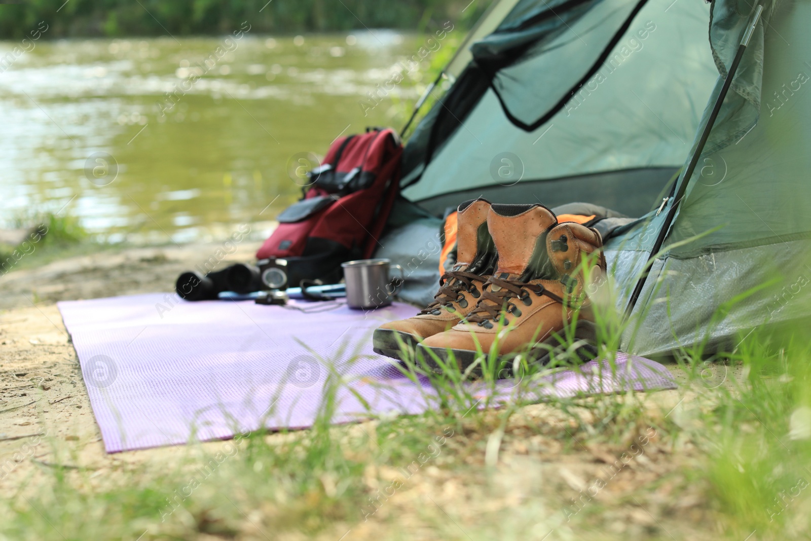 Photo of Boots and camping equipment near tent on riverbank. Space for text