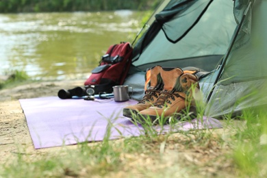 Photo of Boots and camping equipment near tent on riverbank. Space for text