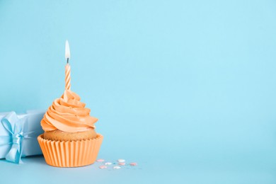 Delicious birthday cupcake with orange cream and burning candle, sprinkles and gift box on light blue background, space for text