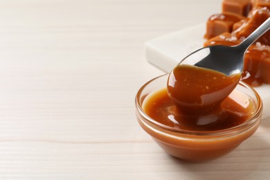 Taking tasty salted caramel with spoon from glass bowl at white wooden table, closeup. Space for text