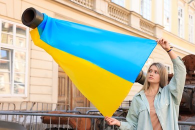 Sad woman holding Ukrainian flag near broken tank in city