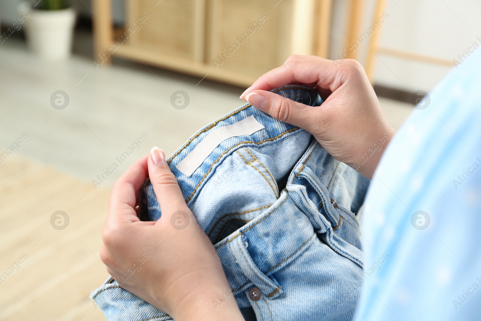 Photo of Woman holding jeans with blank clothing label at home, closeup