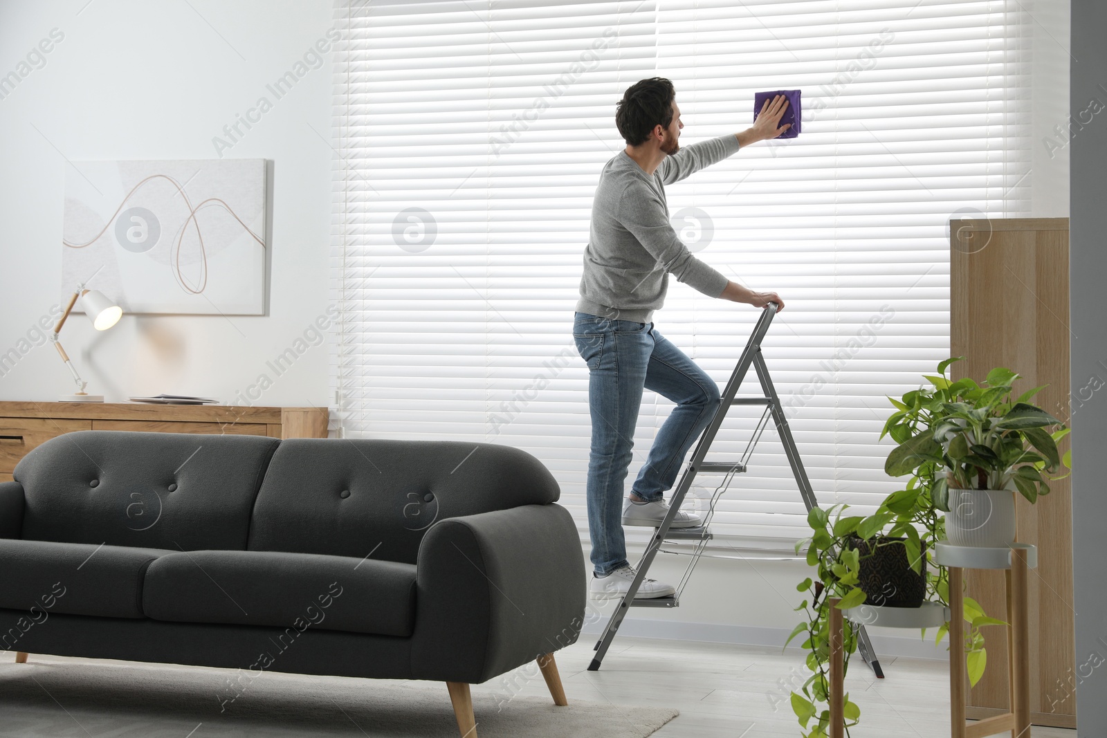 Photo of Man on metal ladder wiping blinds at home