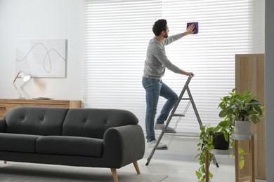 Man on metal ladder wiping blinds at home