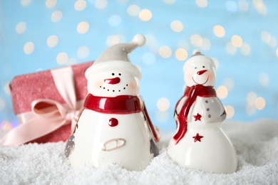 Decorative snowmen near gift box on artificial snow against blurred festive lights