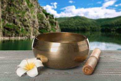 Image of Golden singing bowl, mallet and flower on grey wooden table against mountain landscape 