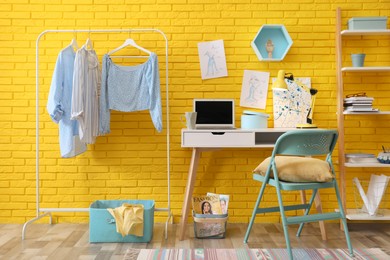 Photo of Fashion designer's workplace with wooden furniture and laptop near yellow brick wall. Stylish interior