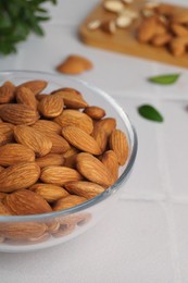 Photo of Bowl of delicious almonds and fresh leaves on white tiled table, closeup