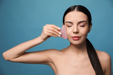 Beautiful young woman doing facial massage with gua sha tool on blue background