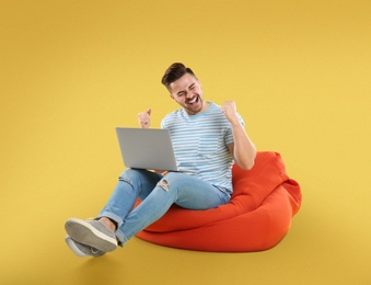 Image of Happy young man with laptop on bean bag chair, yellow background