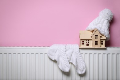 Photo of Modern radiator with knitted hat, socks and wooden house near pink wall indoors, space for text. Winter heating efficiency
