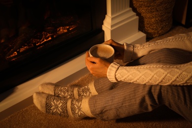 Photo of Woman in warm socks holding hot drink near fireplace with burning wood at home, closeup