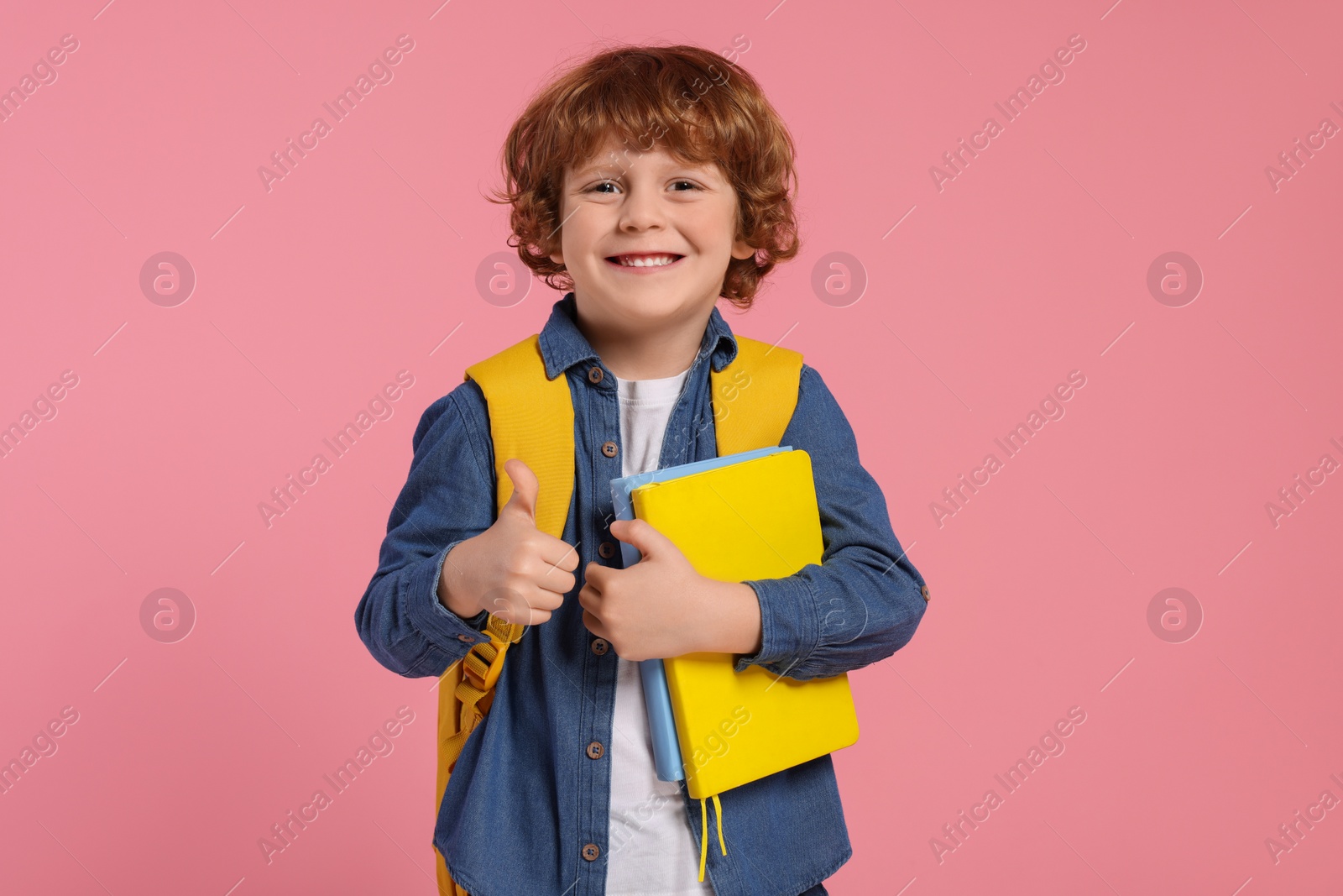 Photo of Happy schoolboy with backpack and books showing thumb up gesture on pink background