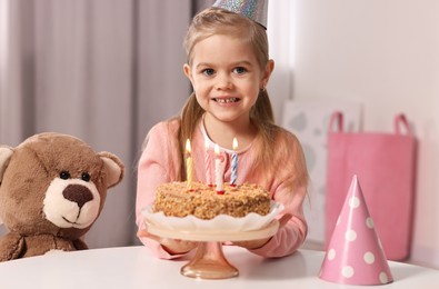 Photo of Cute girl in party hat with birthday cake at table indoors