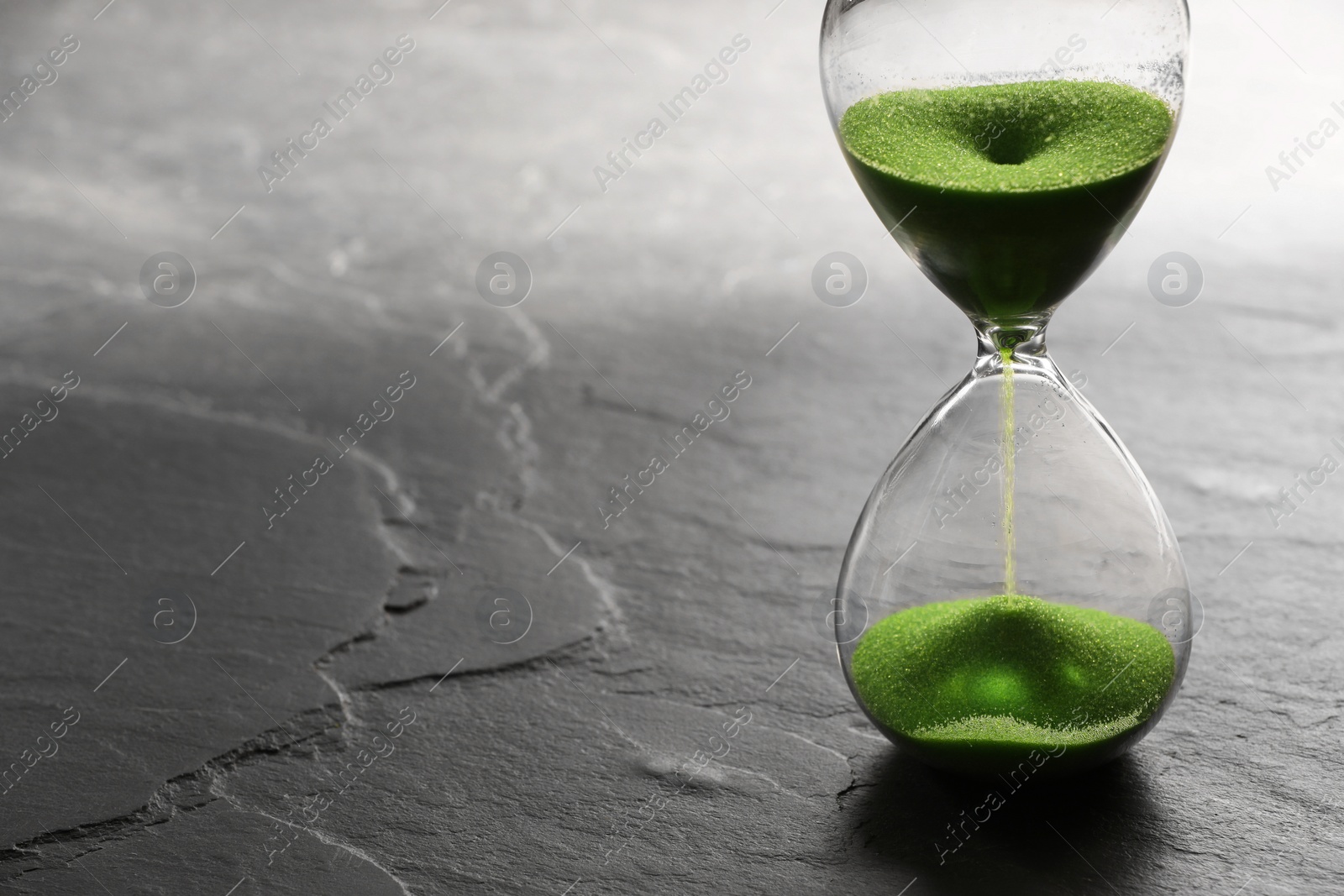 Photo of Hourglass with light green flowing sand on black table. Space for text