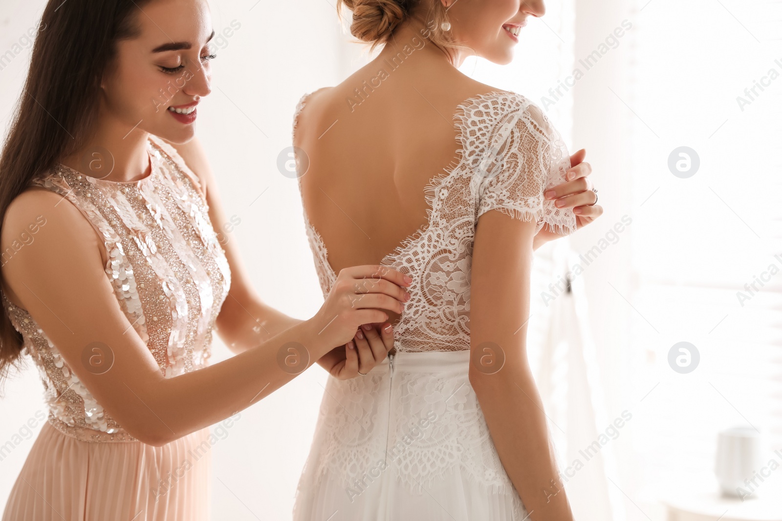 Photo of Young woman helping bride to put on wedding dress in room