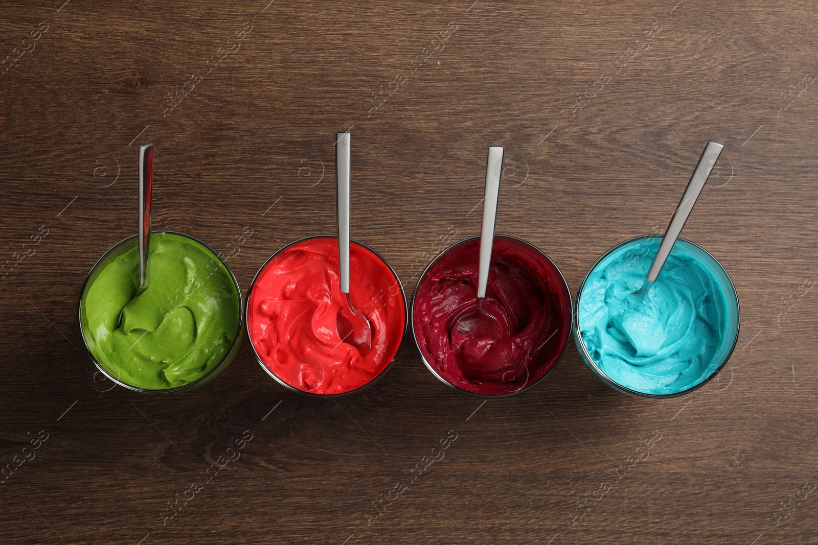 Photo of Glasses of different cream with food coloring on wooden table, flat lay