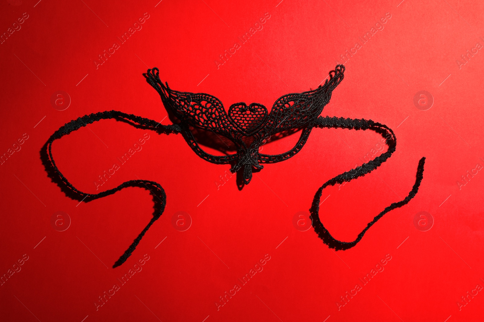 Photo of Black lace mask on red background, top view. Accessory for sexual role play