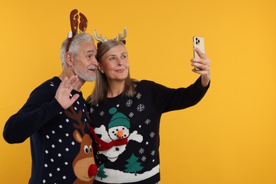 Photo of Senior couple in Christmas sweaters and reindeer headbands taking selfie on orange background. Space for text