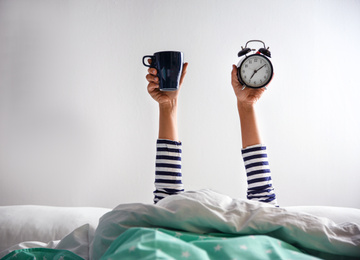 Woman with cup and alarm clock lying in bed, closeup. Morning time