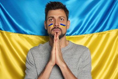 Photo of Sad man with clasped hands praying near Ukrainian flag
