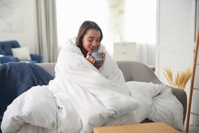 Photo of Beautiful young woman wrapped with soft blanket holding cup of hot drink on sofa at home