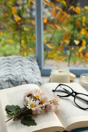 Photo of Open book with chamomile flowers as bookmark, scented candles and glasses on table near window, closeup. Space for text