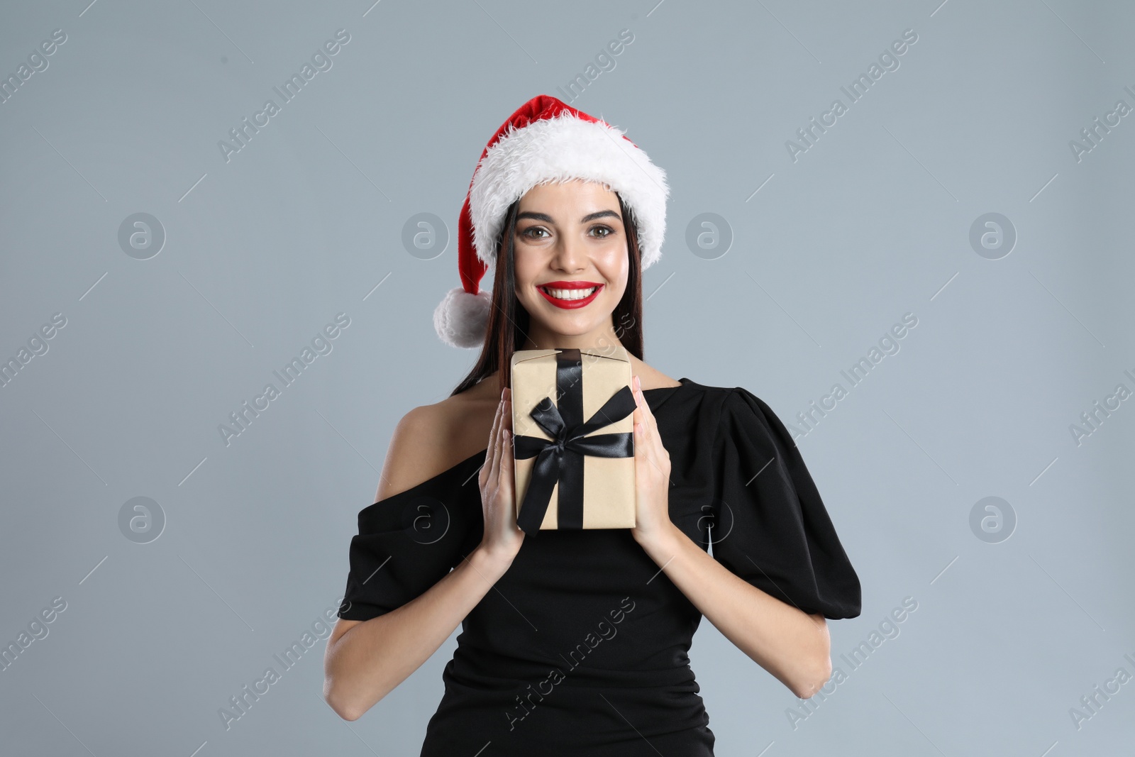 Photo of Woman in black dress and Santa hat holding Christmas gift on grey background