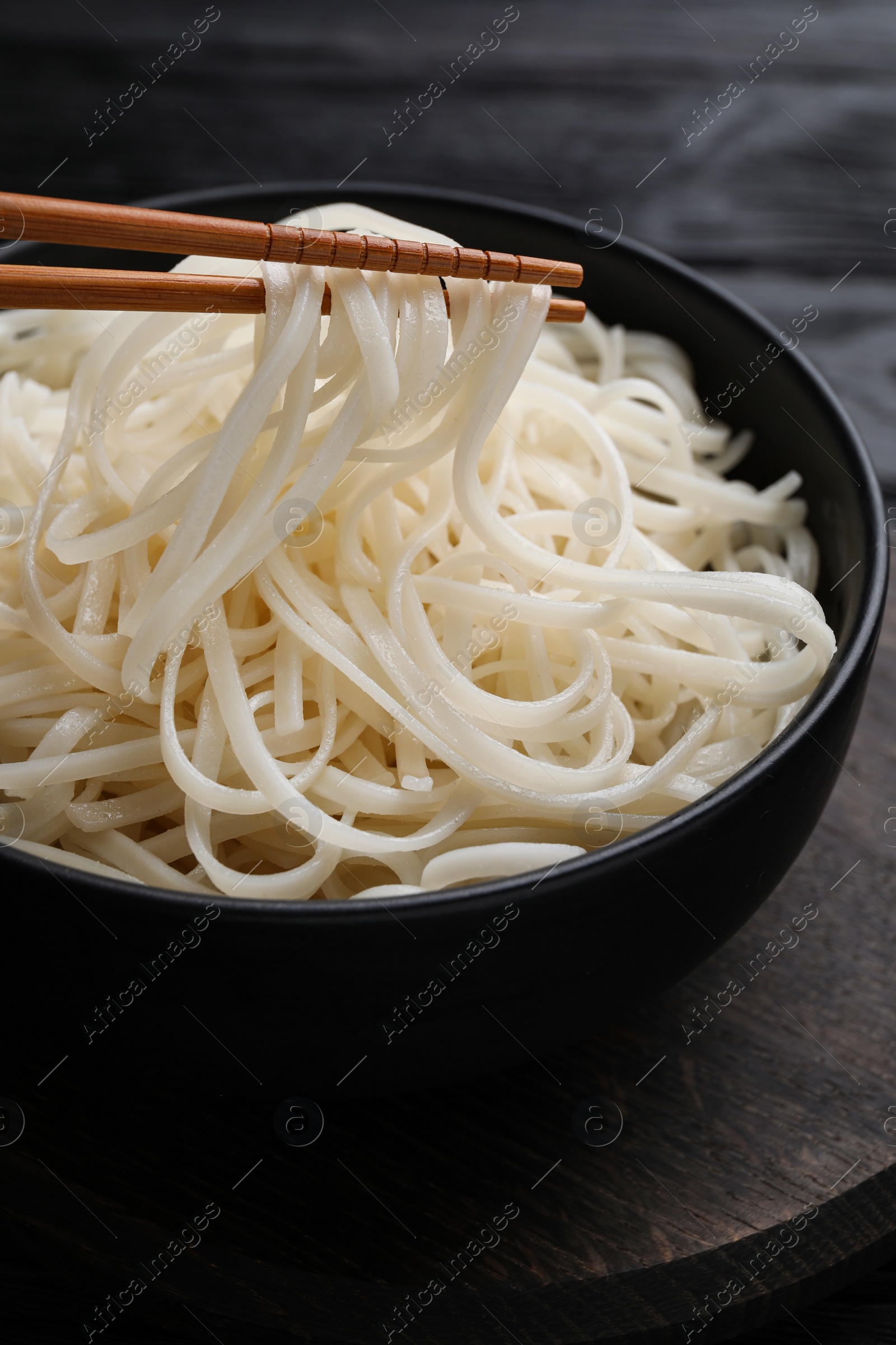 Photo of Taking rice noodles with chopsticks from bowl at black table, closeup