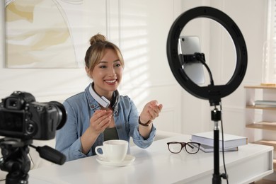 Blogger with cup of tea recording video at table indoors. Using ring lamp and camera